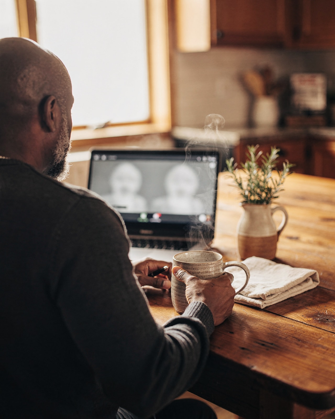 Over-the-shoulder view of a man at a kitchen table on a blurred video consult, morning window light behind.
