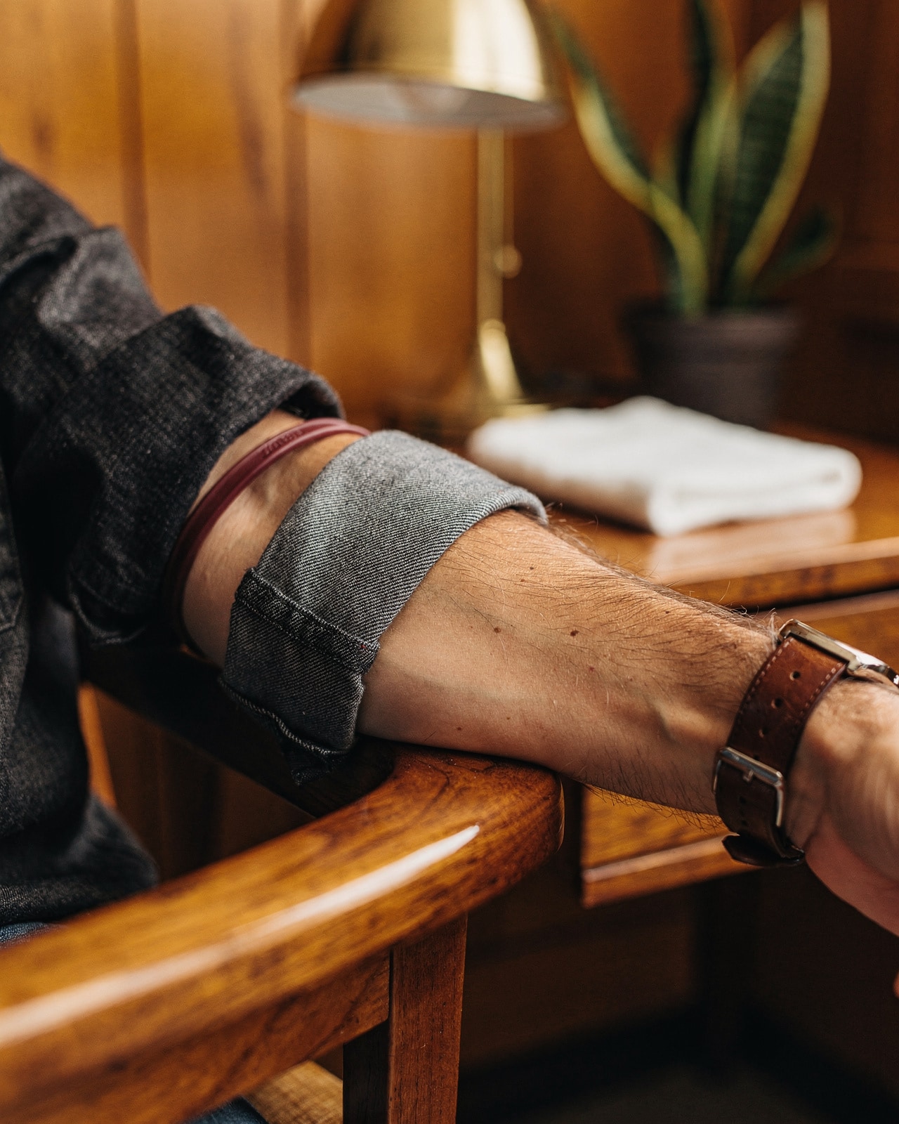 A rolled chambray sleeve on a forearm resting on a warm-wood clinic chair with a tourniquet, no needle visible.