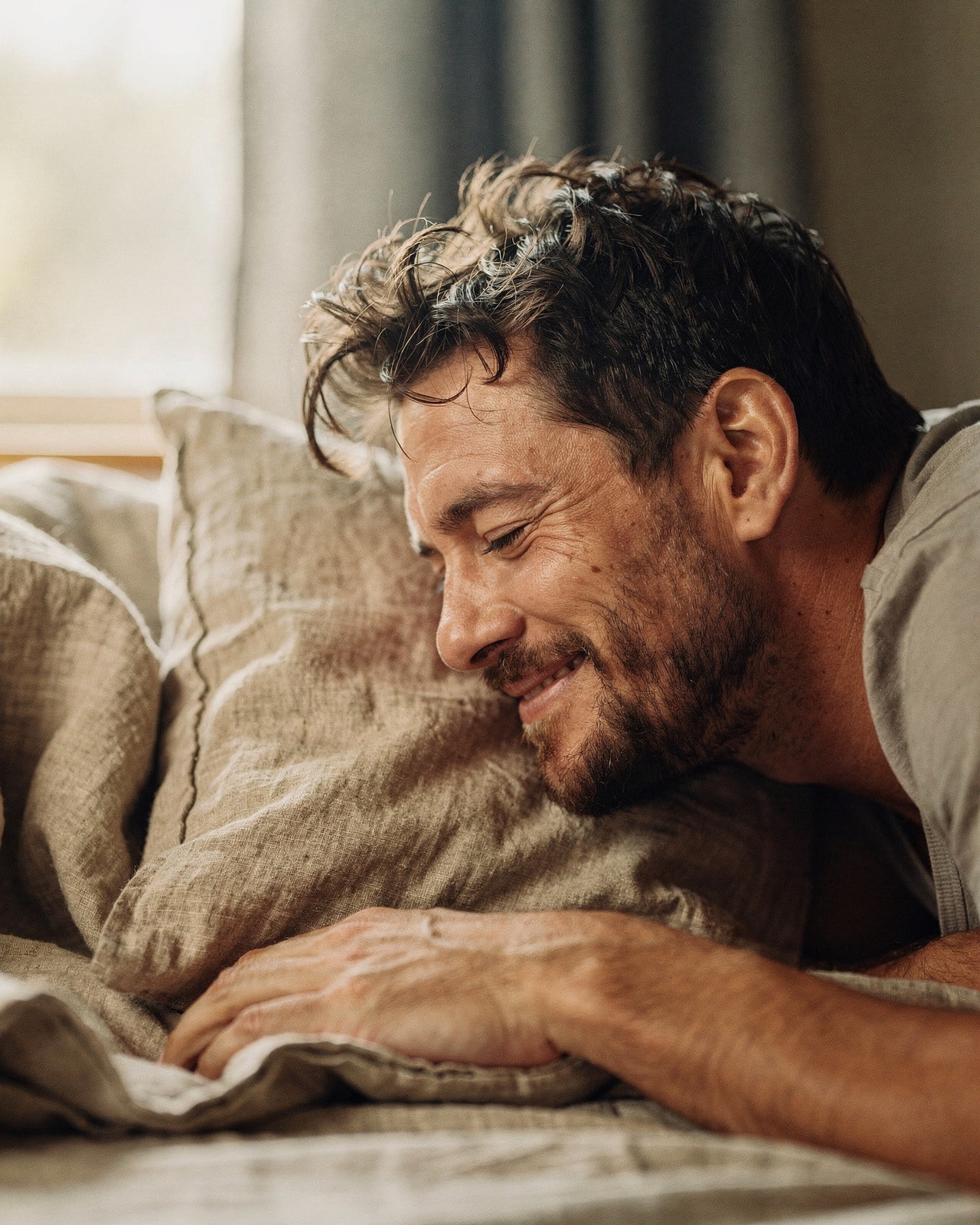 Close-up of a man waking with eyes closed and a small contented smile on an oat-linen pillow in first light.