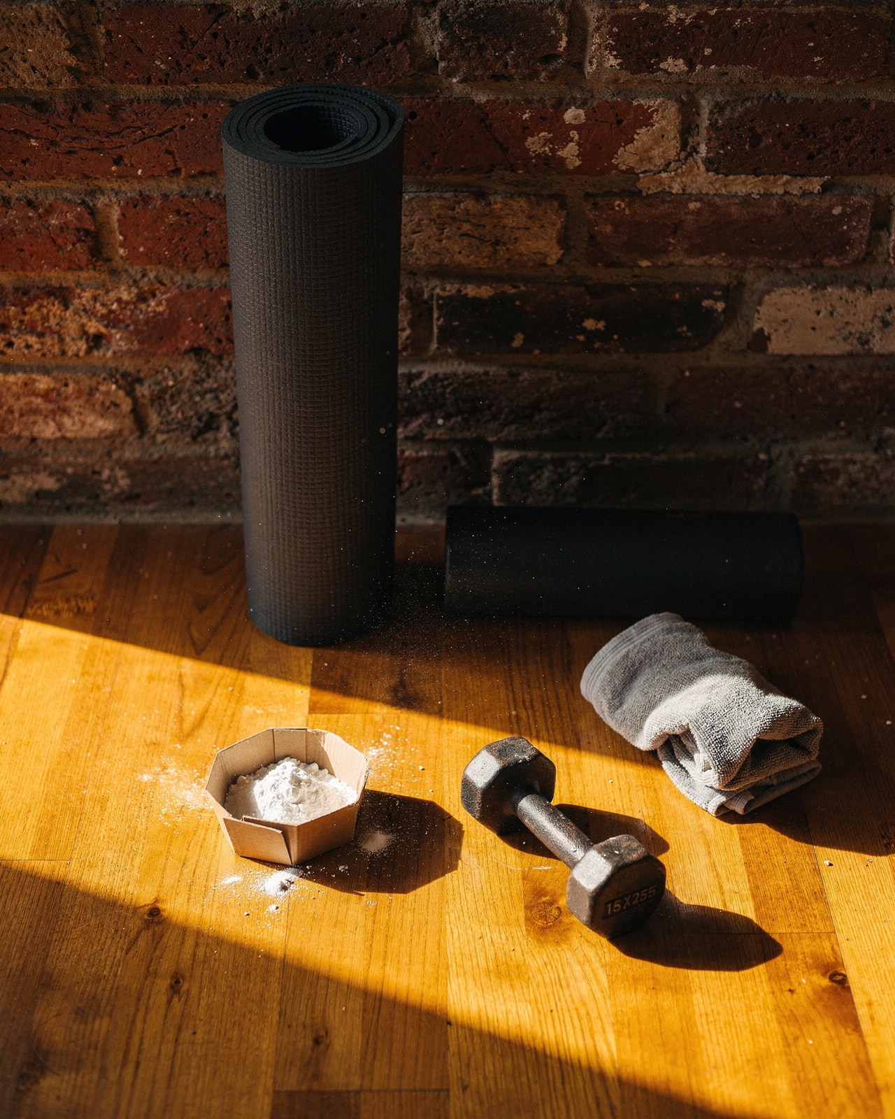 A rolled yoga mat, foam roller, chalk bowl, and dumbbell on a hardwood home-gym floor in warm late afternoon sun.
