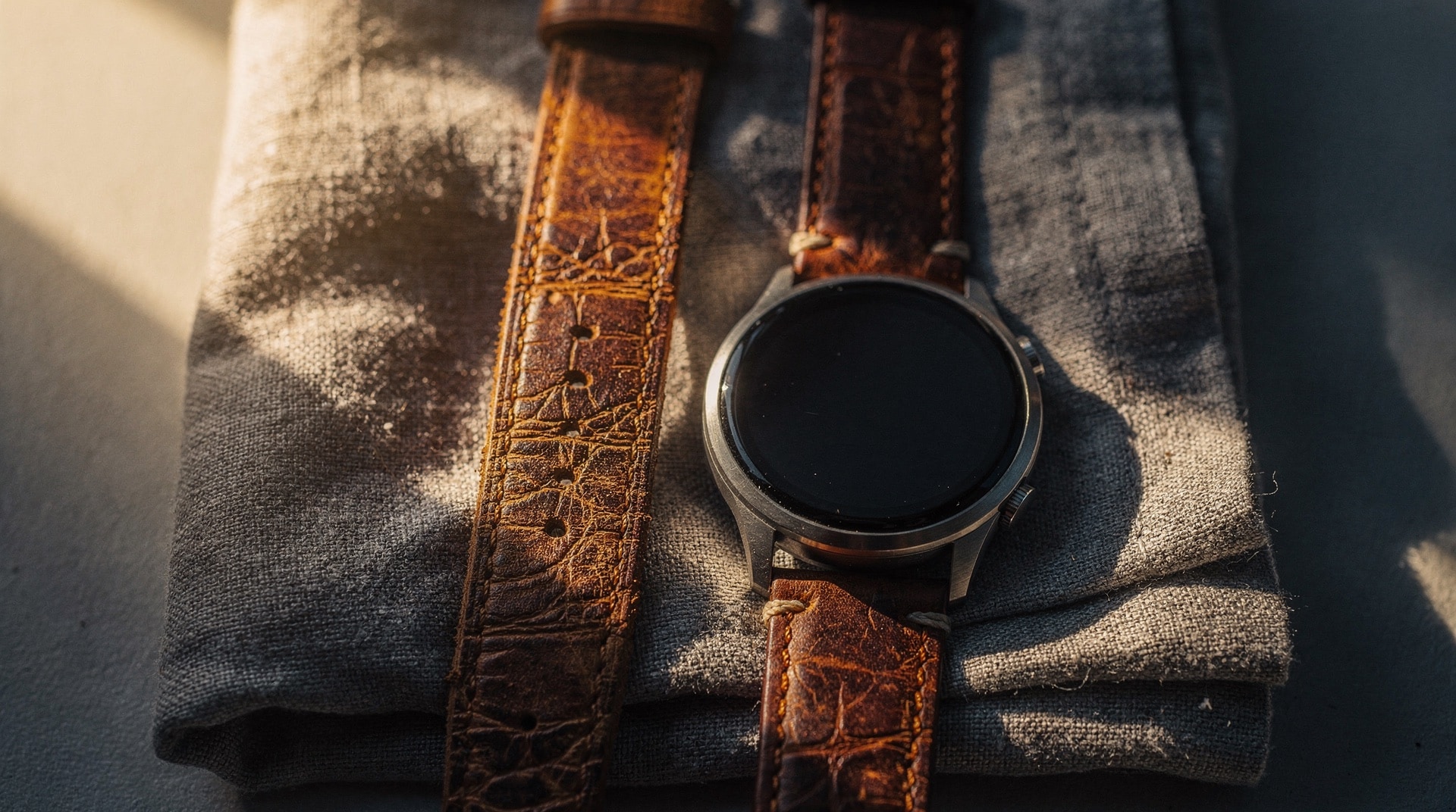 Aged leather watch strap and modern smartwatch resting side by side on folded grey linen at golden hour