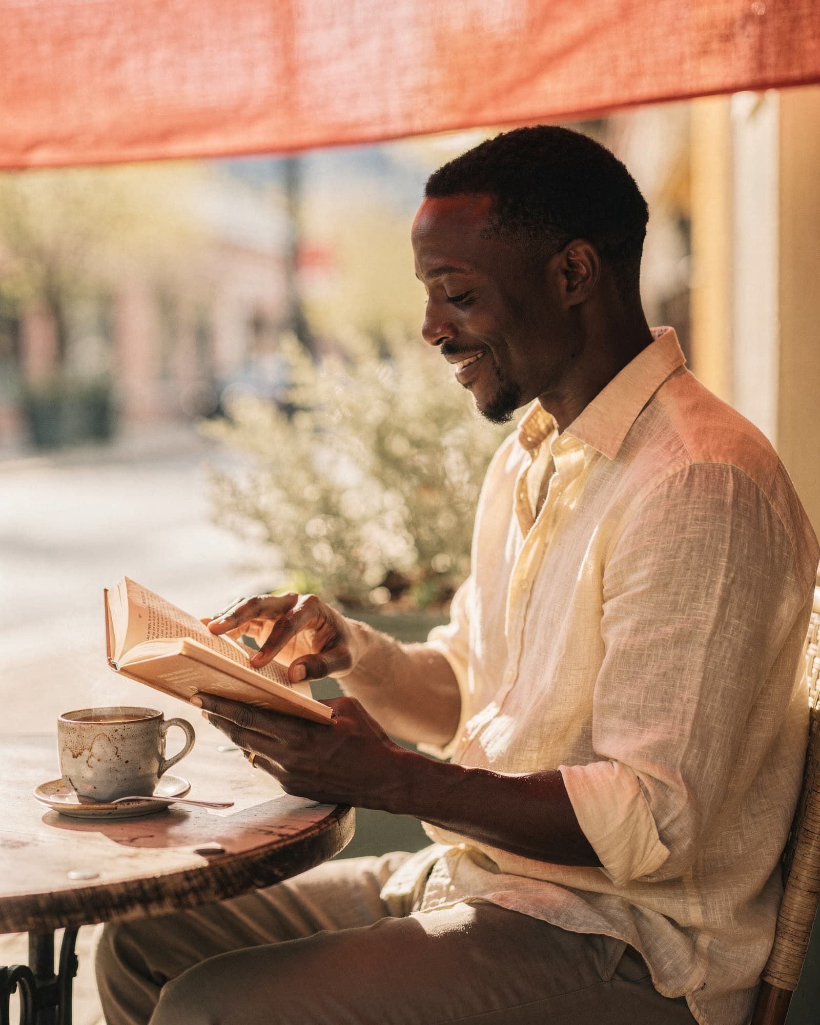 Black man reading a paperback book at a sunlit cafe table on a warm spring afternoon