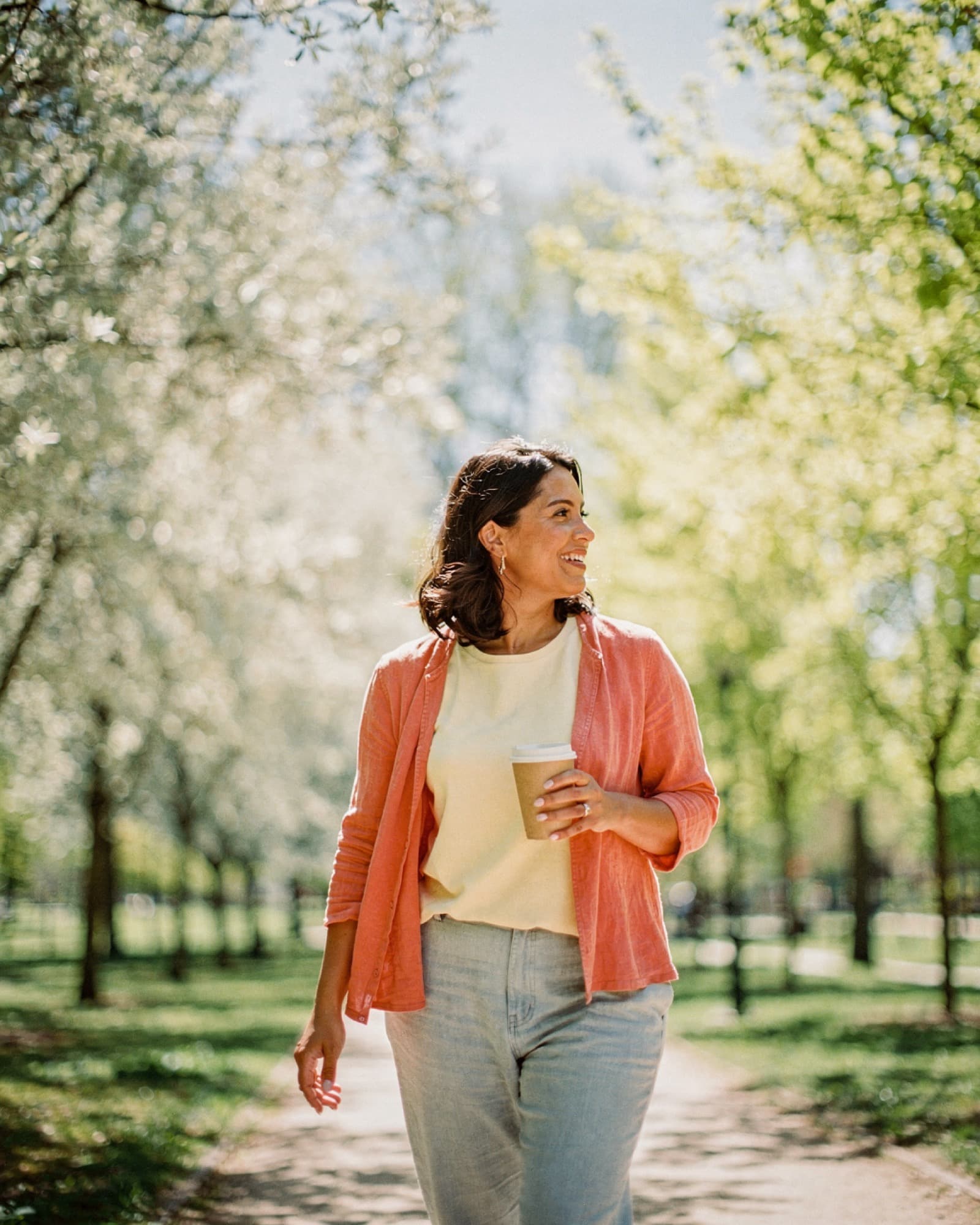 Woman walking on a sunlit park path in late spring, holding a cup of tea