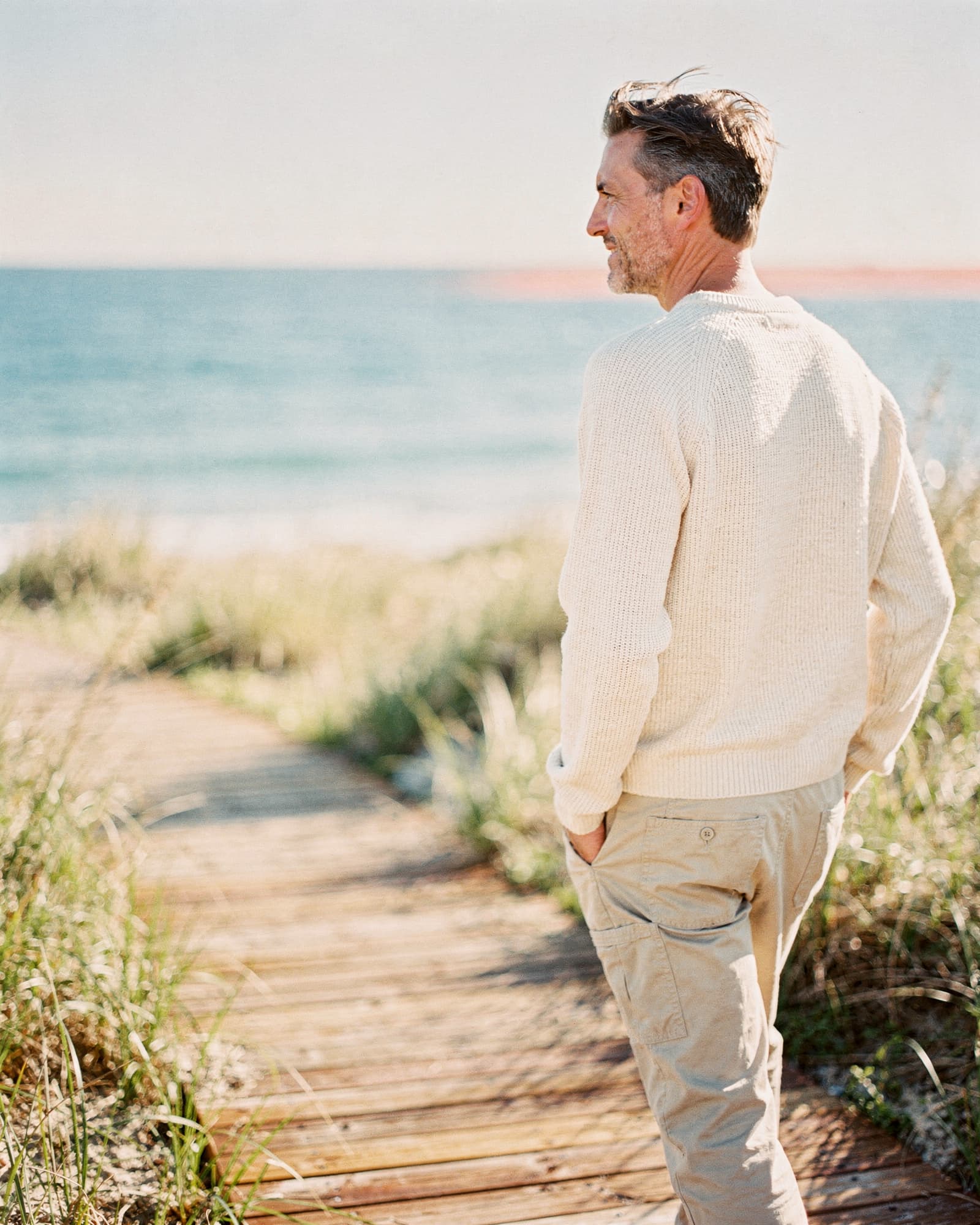Man walking on a coastal boardwalk along sunlit dune grass, looking at the horizon