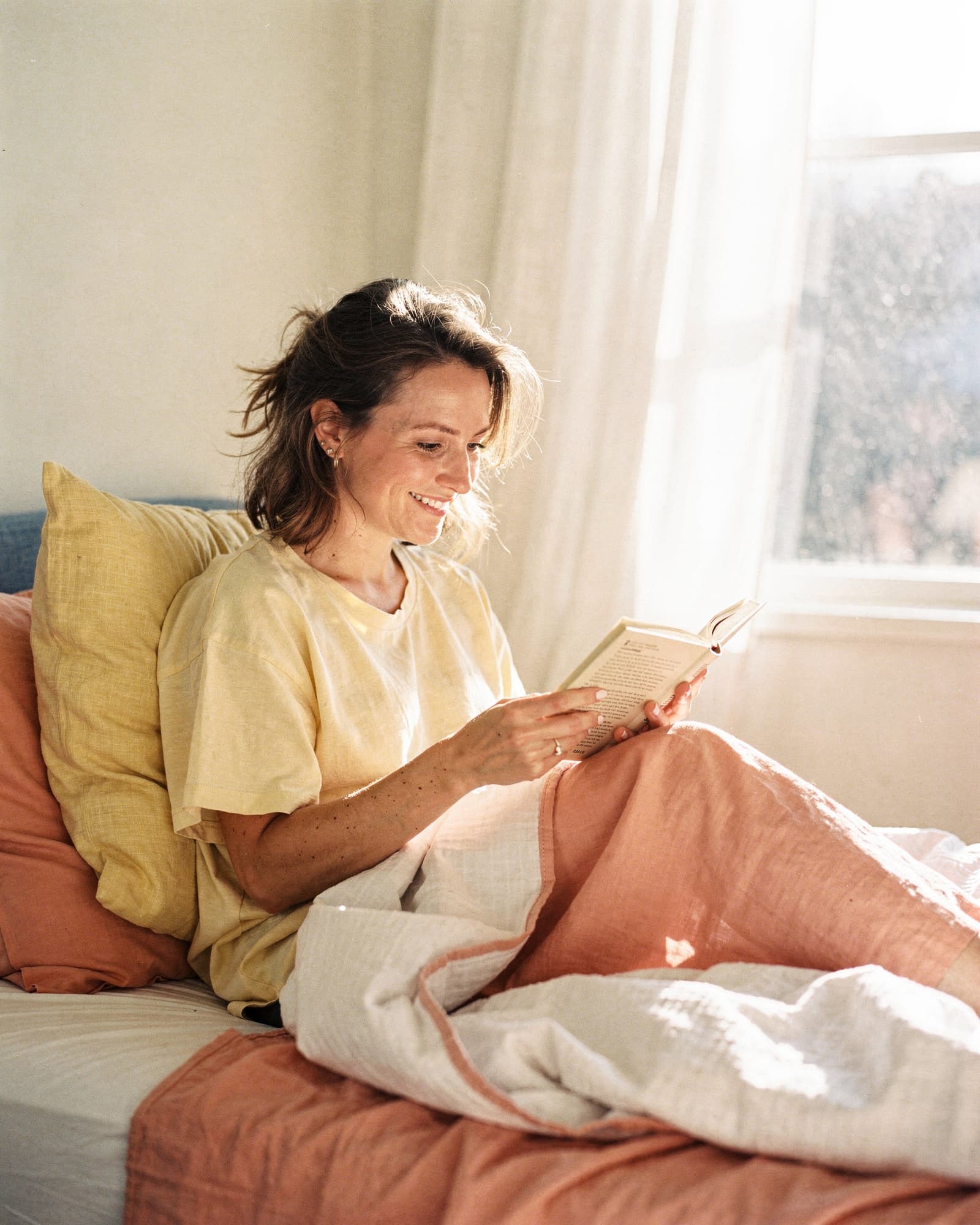 Woman reading a paperback book in a sun-drenched bedroom, soft natural smile